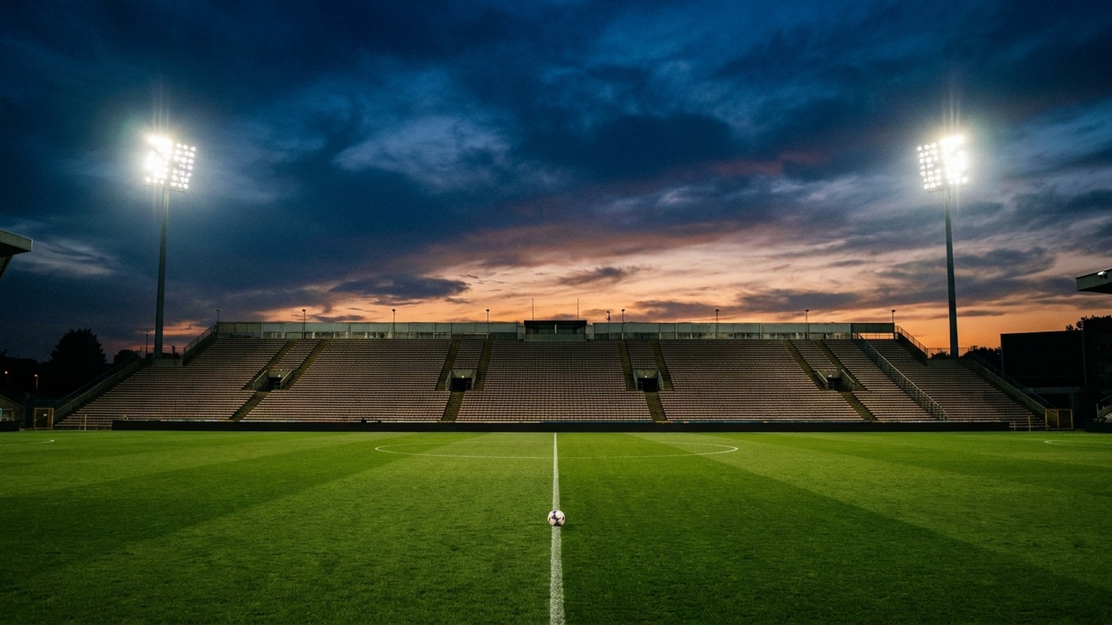 Un grand stade de football à la tombée du jour, tribunes éclairées par des projecteurs, pelouse verte et un ballon posé sur la ligne