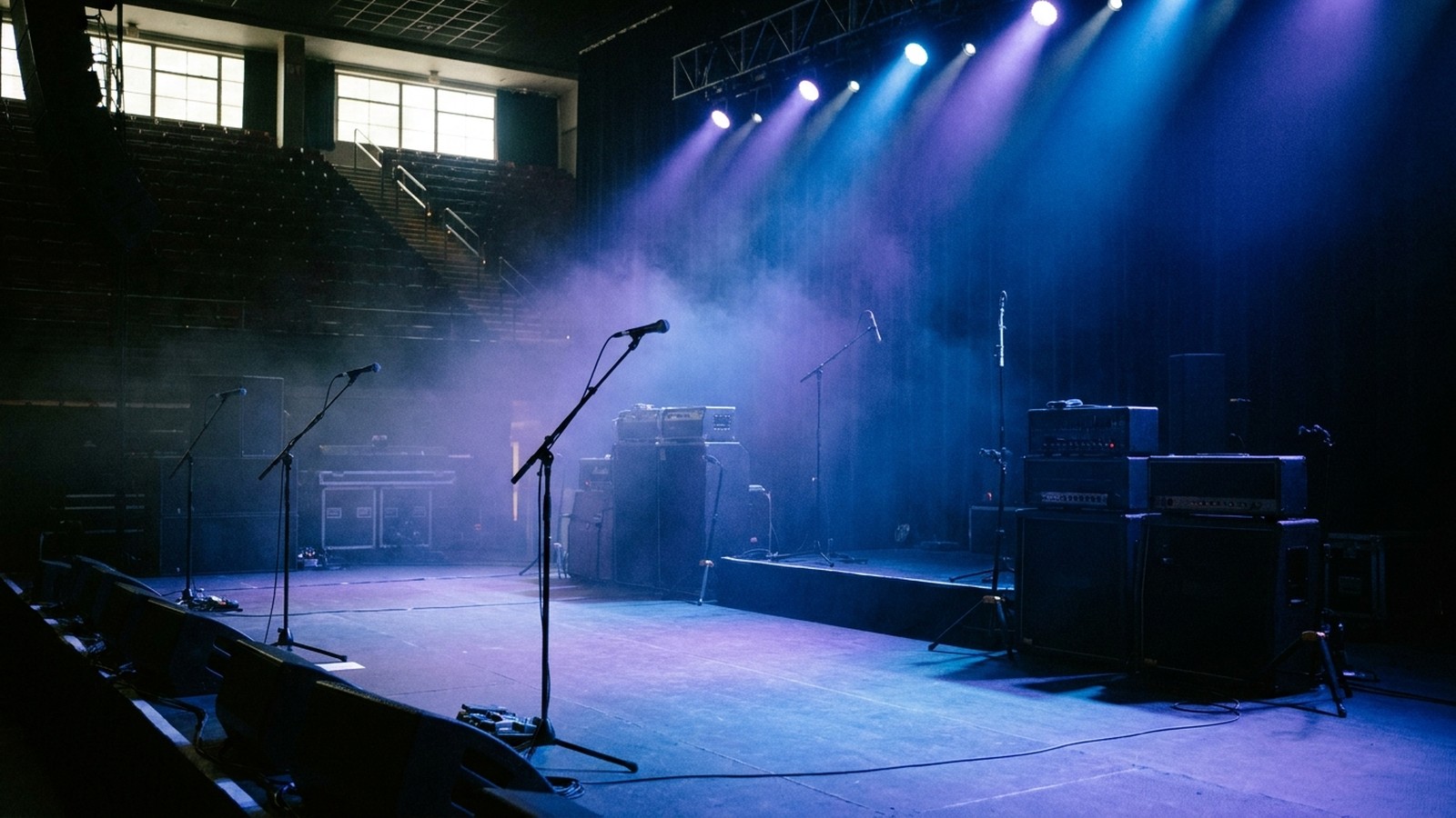 Une scène de concert de rock vide baignée de lumières bleues et violettes, pieds de micro, amplis sombres et fumée, gradins dans l'ombre